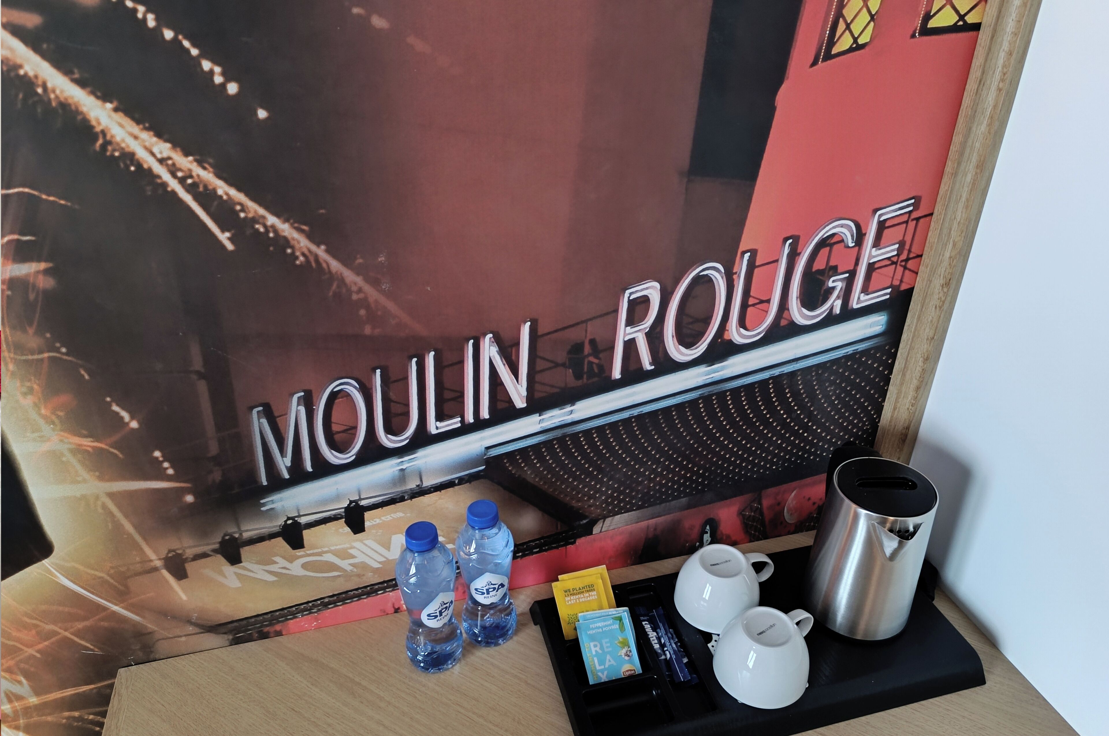 A hotel room with a bed, a nightstand with a lamp, a coffee maker, a water bottle, and a tray with a cup and a box of tissues.