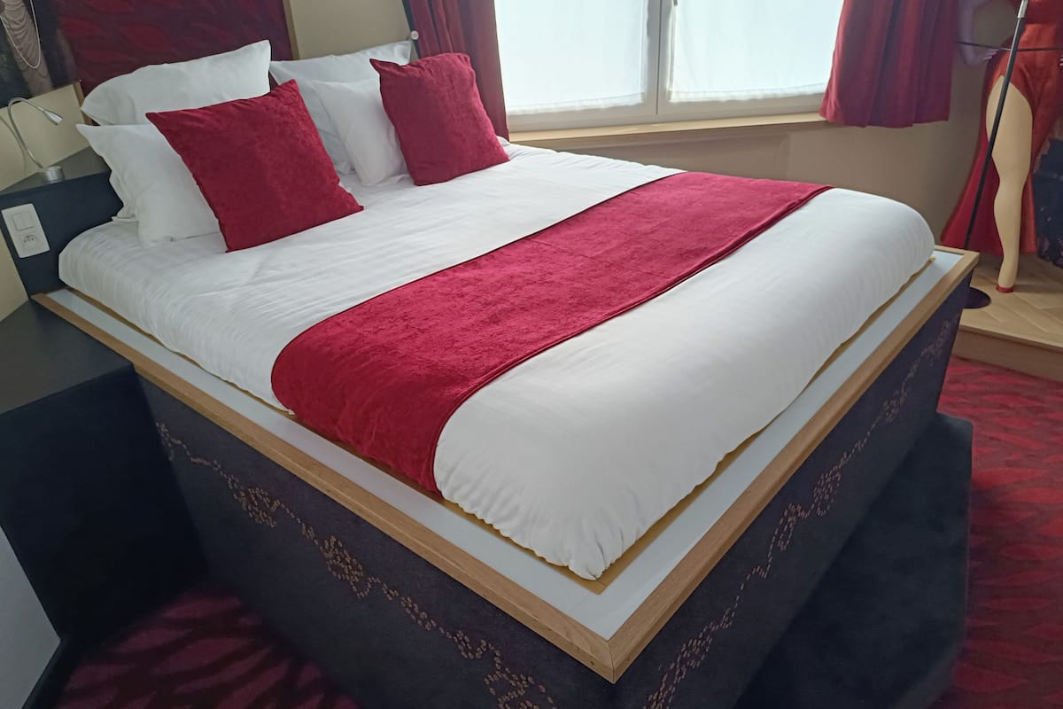 Ein ordentlich bezogenes Bett mit weißer und roter Bettwäsche, ein hölzerner Kopfteil und ein Fenster mit Vorhängen.