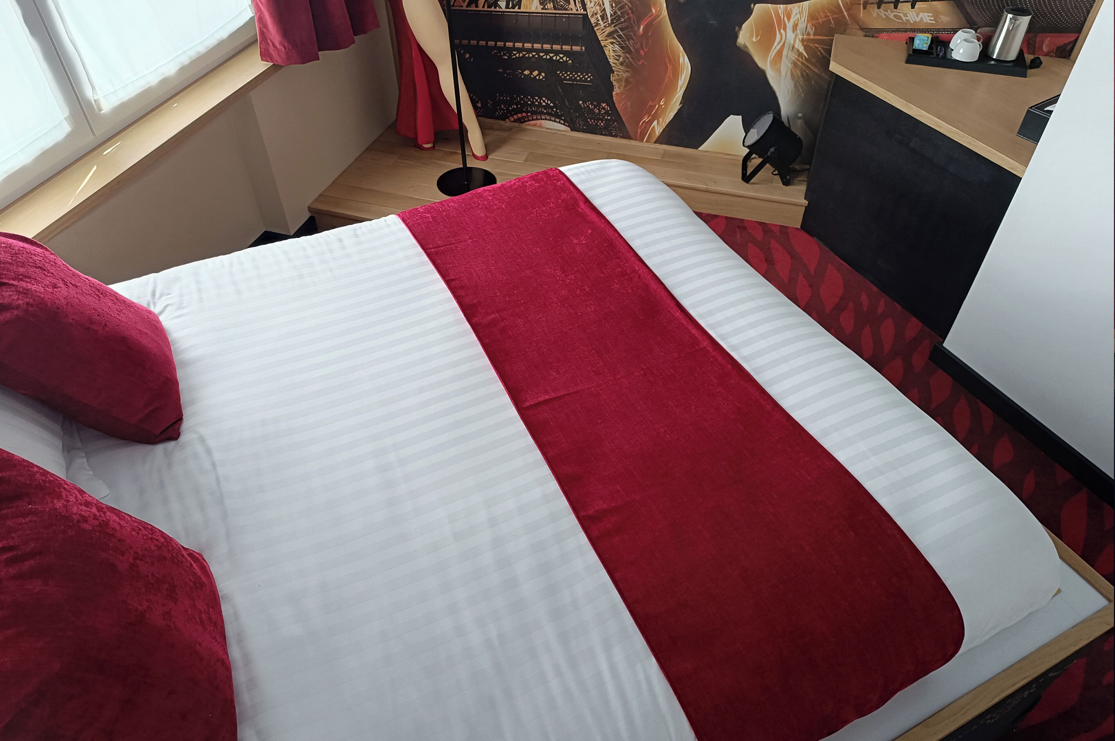 A hotel room with a bed featuring a red and white striped bedspread, a wooden floor, and a window with curtains.