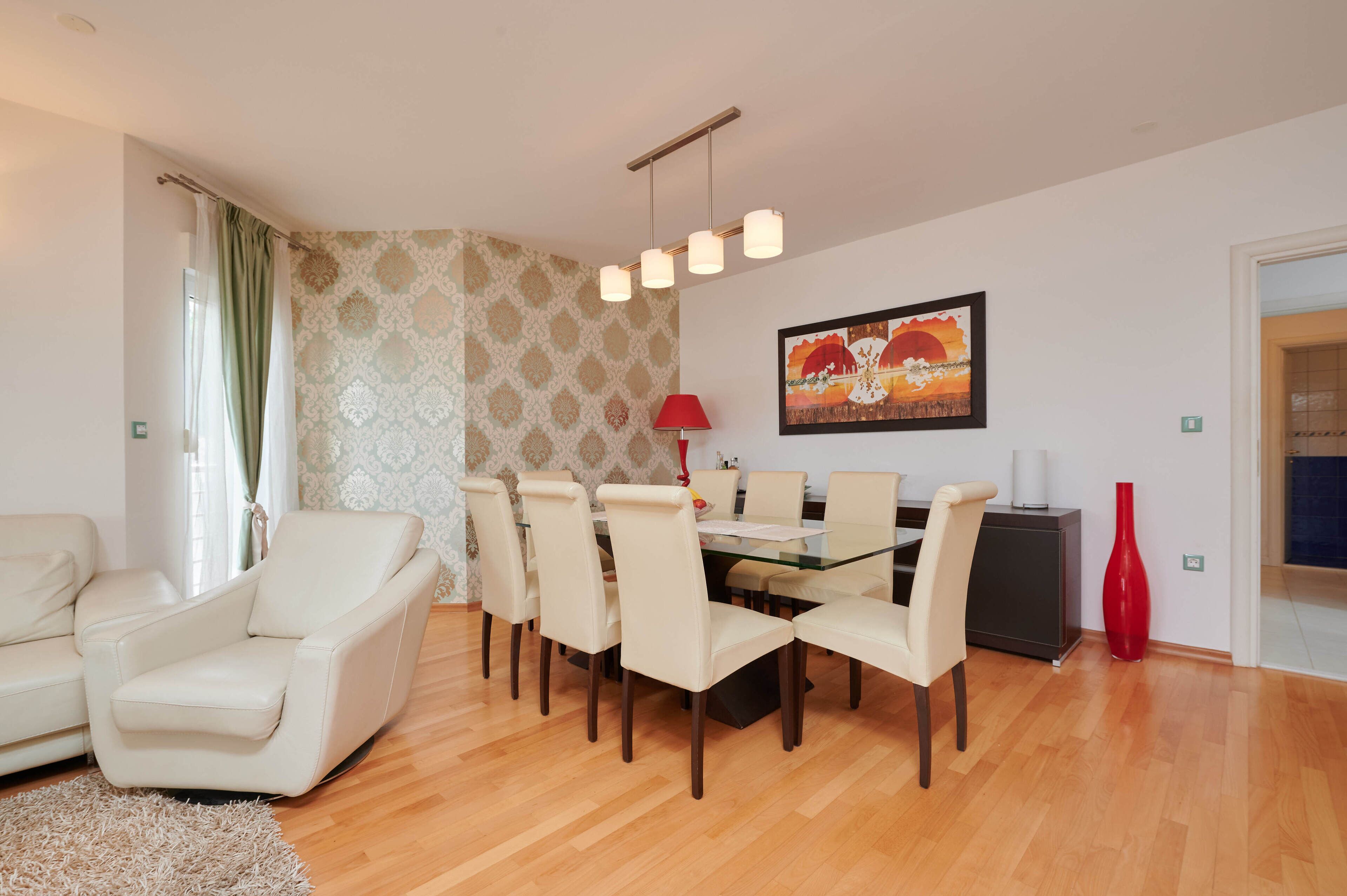 A dining area with a glass table, six chairs, a red lamp, and a painting on the wall.