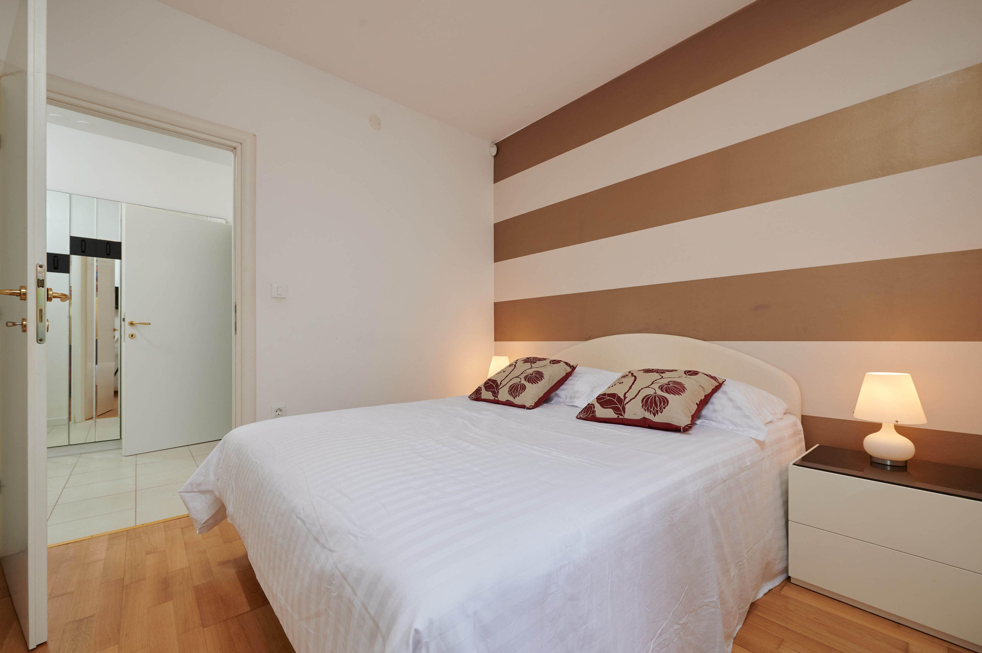 A neatly made bed with white bedding and two decorative pillows, a bedside lamp, and a wooden floor.