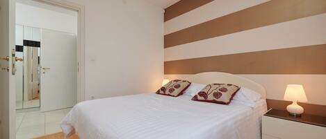 A neatly made bed with white bedding and two decorative pillows, a bedside lamp, and a wooden floor.