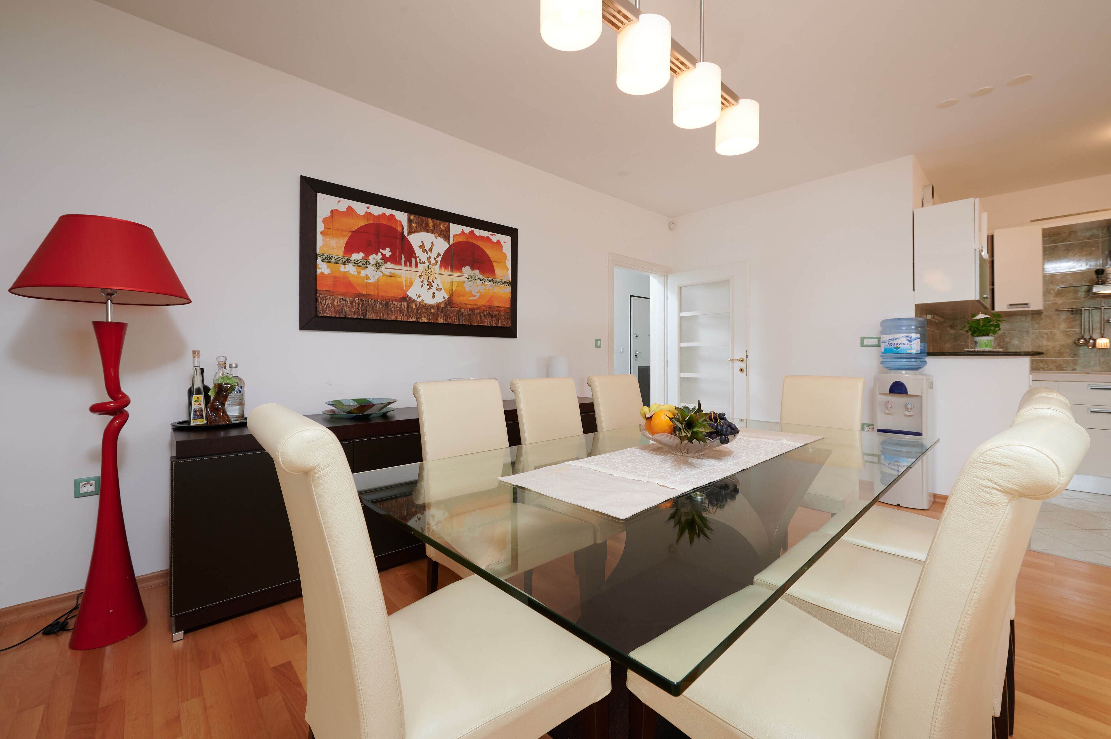 Un espace repas moderne avec une table en verre, des chaises blanches, une lampe rouge, une œuvre d’art encadrée et une cuisine en arrière-plan.