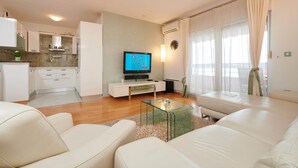 Een moderne woonkamer met een glazen salontafel, witte banken, een televisie waarop een doolspel wordt getoond, een keuken op de achtergrond, en een groot raam met gordijnen.