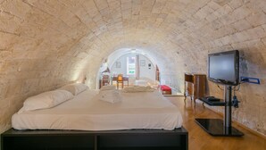 Una habitación con techo abovedado de piedra, cama con ropa blanca, televisor sobre un mueble y escritorio de madera con una silla.
