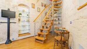 Una escalera con peldaños de madera y barandillas de metal, un televisor de pantalla plana sobre un soporte, y una pequeña mesa con sillas en una habitación con paredes de piedra y piso de madera.