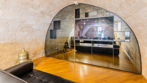 Un interior moderno con un arco de piedra, un espejo grande que refleja una habitación con un sofá y una lámpara decorativa.