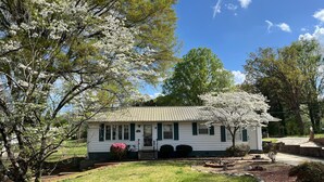 A white single-story house with a green roof, surrounded by a well-maintained lawn and flowering trees.