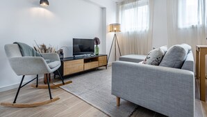 Een moderne woonkamer met een grijze bank, een schommelstoel, een televisie op een houten kast en een vloerlamp.