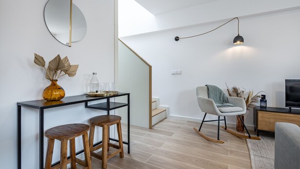 Een moderne woonkamer met een houten salontafel, twee houten krukken, een ronde spiegel, een bank, een televisie en een trap.