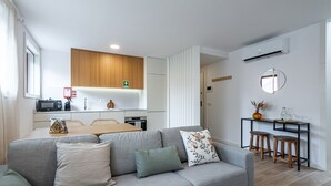 Een moderne woonkamer met een grijze bank, een eettafel en een keukenhoek met houten kastjes en witte apparaten.