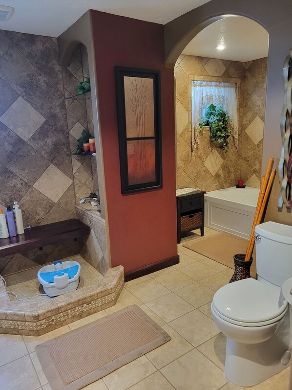 A bathroom with a toilet, bidet, and bathtub. There is a shelf with toiletries and a framed artwork on the wall.