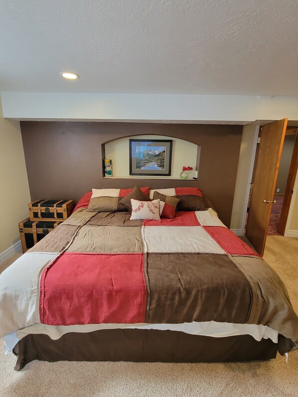 Un lit bien fait, recouvert d’une couverture rouge et marron, avec un tableau encadré au-dessus et une commode en bois sur le côté gauche.