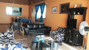 Una sala de estar con un sofá de cuero negro, un sofá estampado, una mesa de centro de vidrio, una silla de ruedas, un ventilador y un televisor.