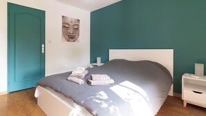 A bedroom with a bed, a teal wall, a wooden floor, a white nightstand, and a door. - Jolie Villa Liwa Entièrement Rénovée Avec Piscine - 8 Pers (Velaux)