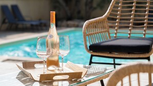 A glass table with a bottle and two glasses, a wicker chair with a cushion, and a pool in the background. - Jolie Villa Liwa Entièrement Rénovée Avec Piscine - 8 Pers (Velaux)