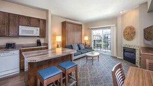 Una cocina moderna con armarios de madera, un microondas y una zona de comedor con mesa de madera y taburetes. Hay un sofá con cojines estampados y una pequeña mesa redonda con una lámpara. Se ve una chimenea en la sala de estar, y un balcón con muebles de exterior a través de las puertas de cristal.