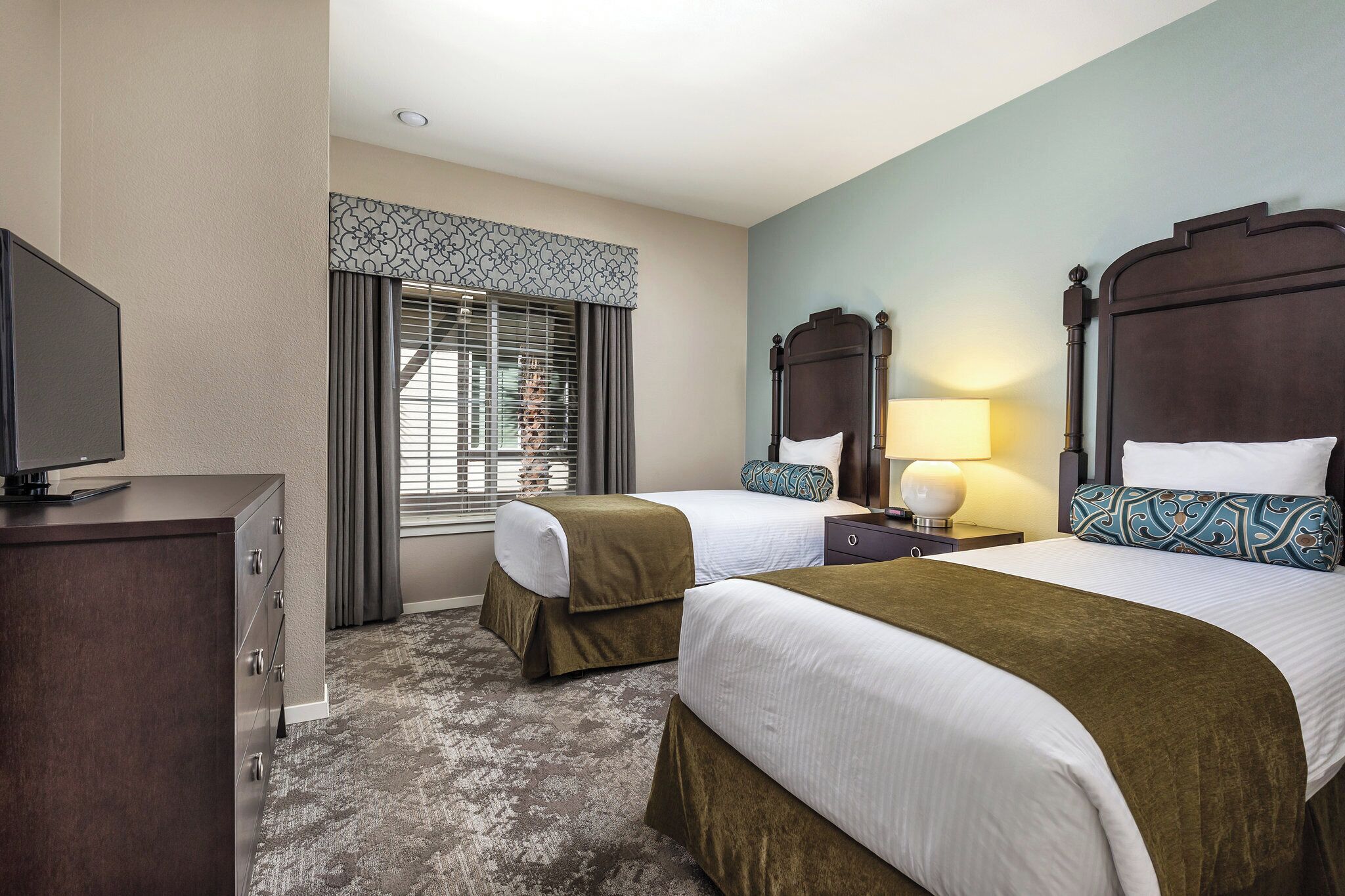 A hotel room with two beds, a TV, a desk, and a window with curtains.