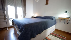 A bedroom with a bed, a nightstand with a lamp, a window with blinds, and a door.