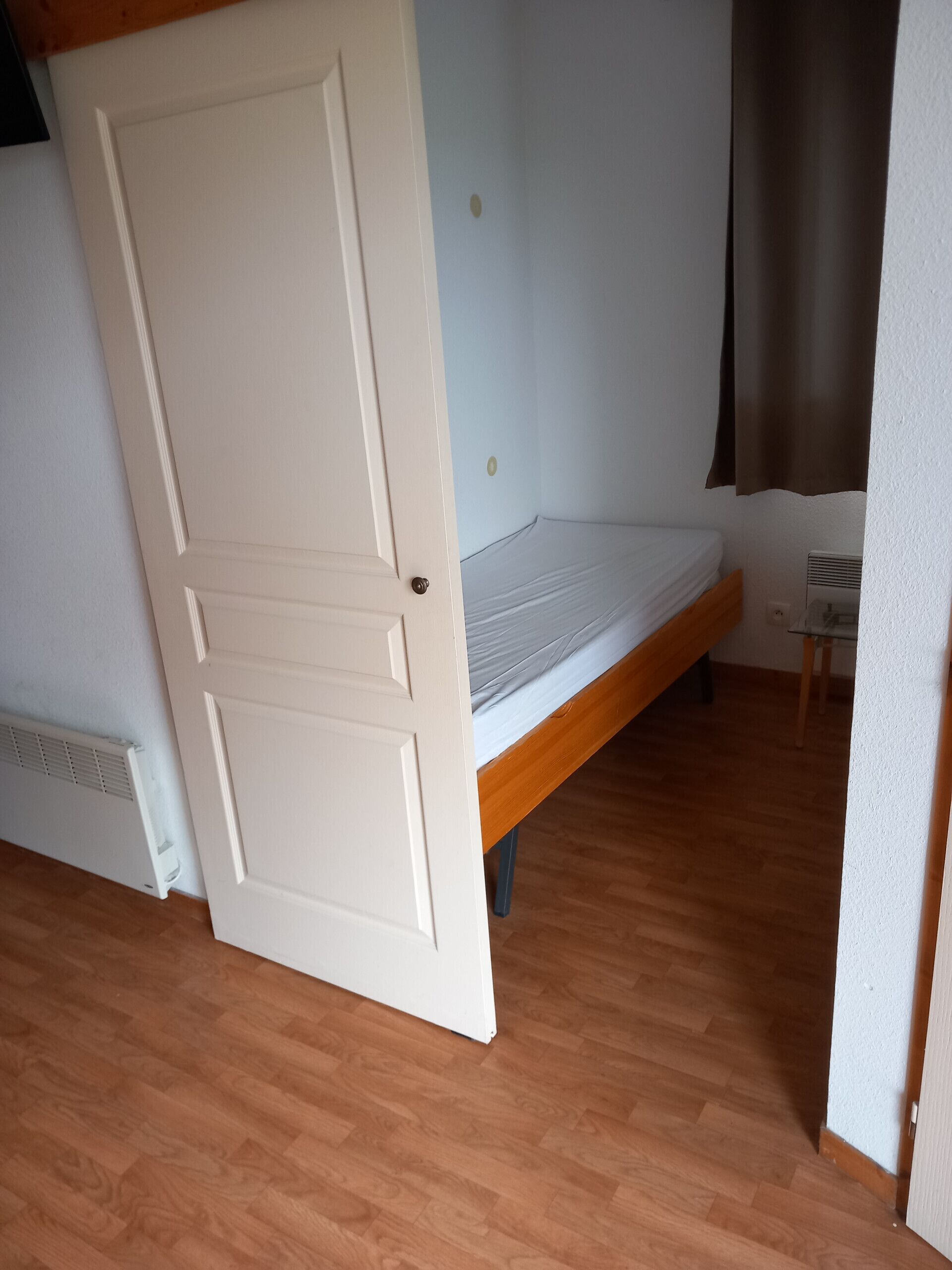 A room with a wooden floor, a bed with a white headboard, a radiator, and a white door.