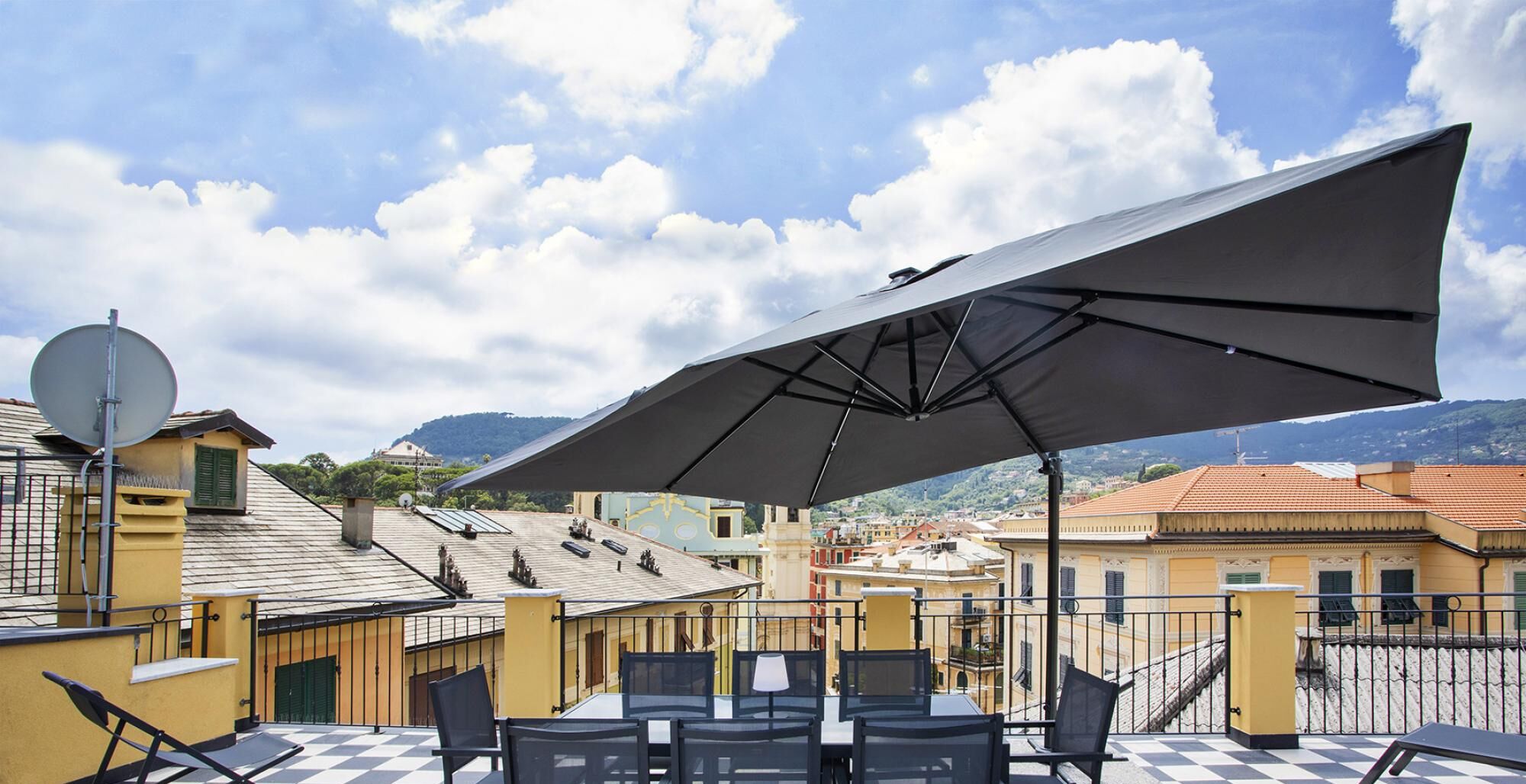 An outdoor terrace with tables and chairs, a large umbrella, and a view of buildings and mountains.