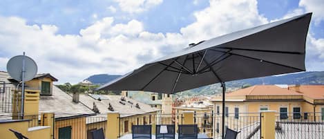Una terraza al aire libre con mesas y sillas, un paraguas grande y vistas a edificios y montañas.