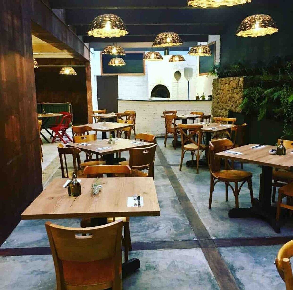 An indoor dining area with wooden tables and chairs, hanging pendant lights, and a stone wall in the background.