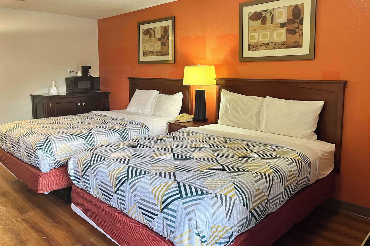 A hotel room with two beds, each with patterned bedding and pillows, a nightstand with a lamp, a telephone, and a small table with a coffee maker.