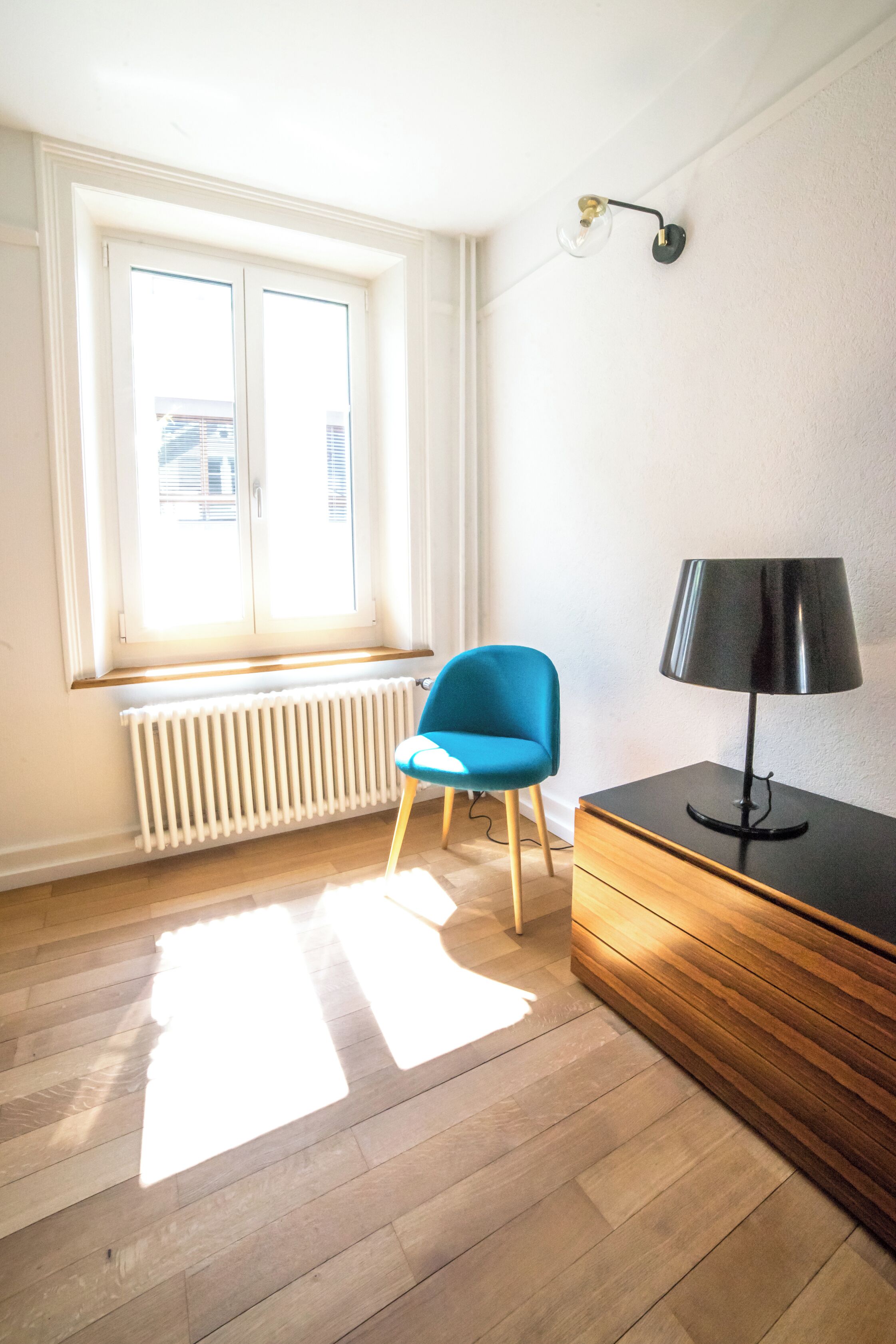 A room with a window, a radiator, a blue chair, a black lamp, and a wooden cabinet.