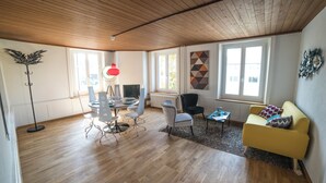 A modern living room with a wooden ceiling, a yellow sofa, a dining table with chairs, and a floor lamp. - Tilia Apartments (Uster)