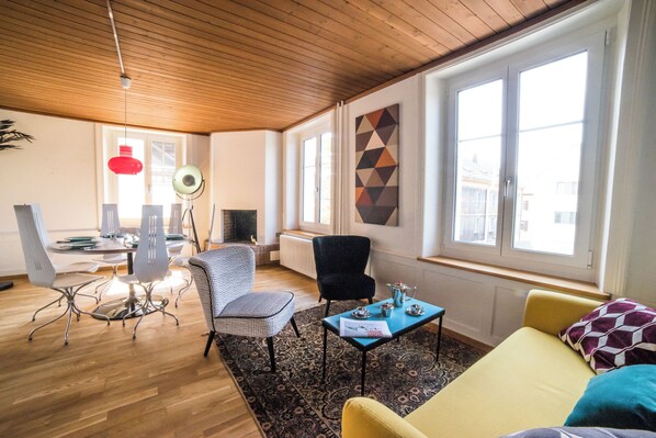 A modern living room with a wooden ceiling, a dining area with a table and chairs, a sofa, a patterned rug, and large windows with a geometric wall decoration.