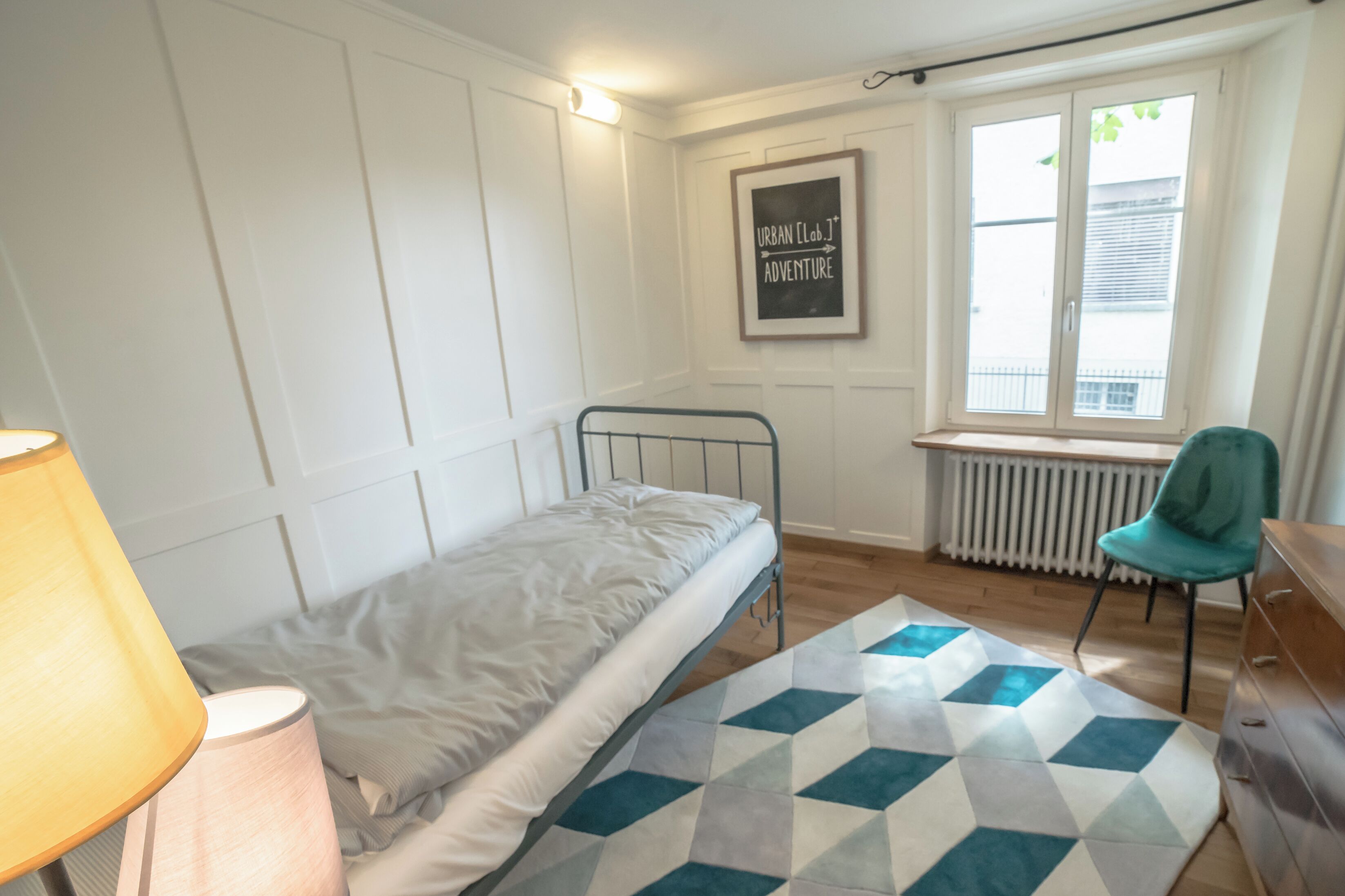 A small, neatly arranged bedroom with a single bed, a bedside lamp, a radiator, a window with blinds, and a framed motivational poster on the wall.