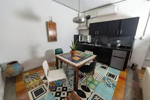 Una cocina compacta con armarios negros, un microondas y un lavamanos. Hay una mesa de comedor con sillas y una planta en maceta. El suelo tiene azulejos con diseños coloridos.