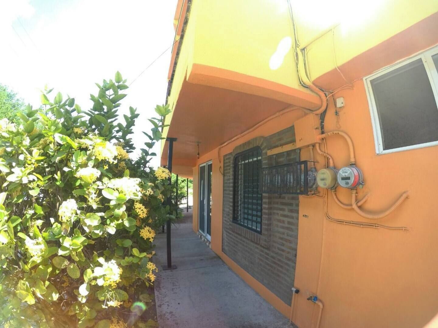 A building with a yellow exterior, a window with a metal grill, and a hose connected to a wall-mounted device.