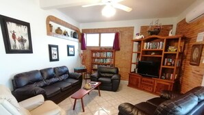 A living room with a sofa, armchair, and a coffee table. There is a bookshelf with books and a television. - 2 BdD Villa Completa Casalaspalmas-punta De Mita (Punta de Mita)