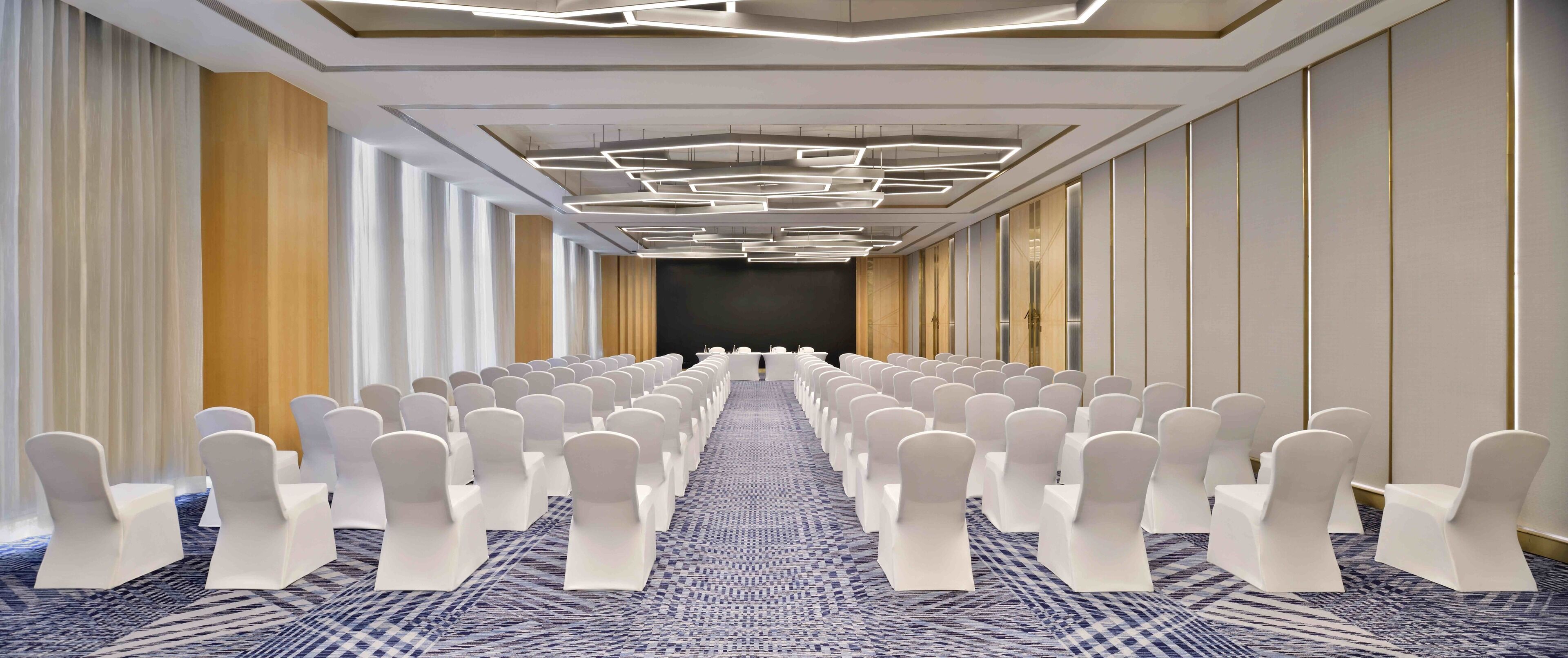 Un salón de conferencias con hileras de sillas blancas, una alfombra estampada y un diseño moderno en el techo.