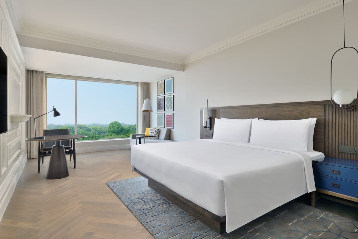 A spacious bedroom with a large bed, a desk with a chair, a blue nightstand, and a large window offering a view of greenery.
