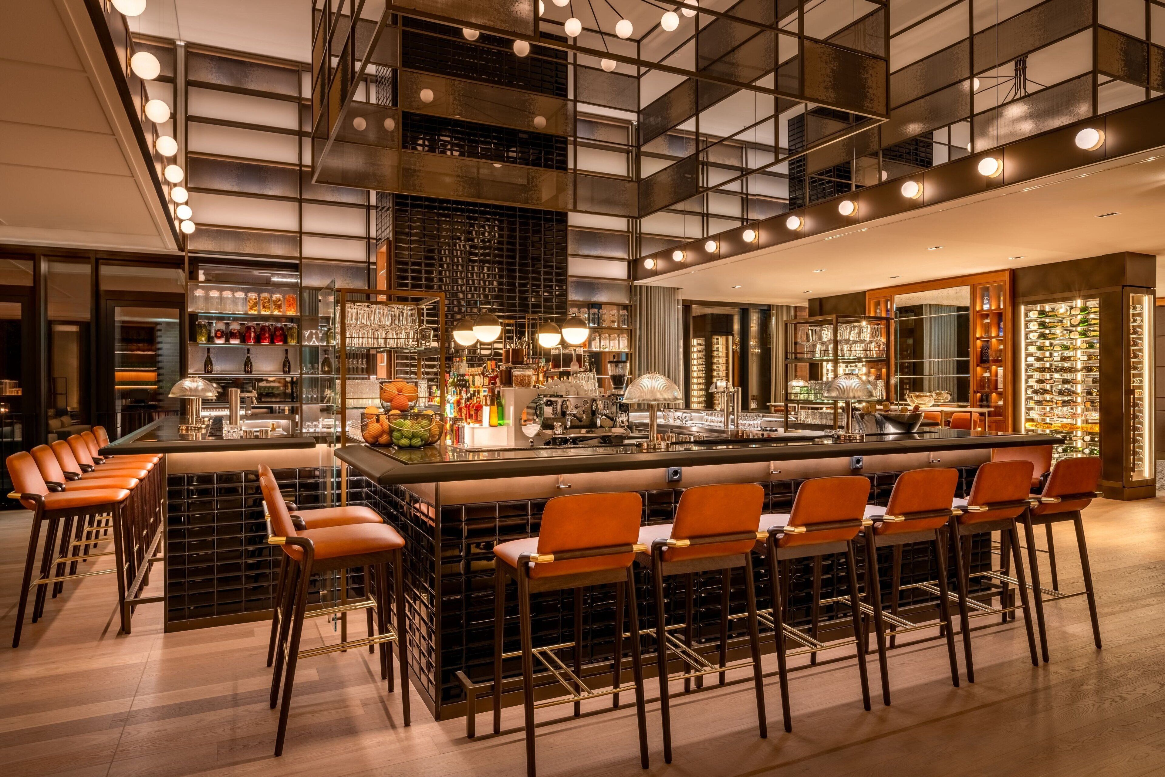 Uma área de bar moderna com um balcão comprido, vários bancos e uma variedade de garrafas e copos nas prateleiras.