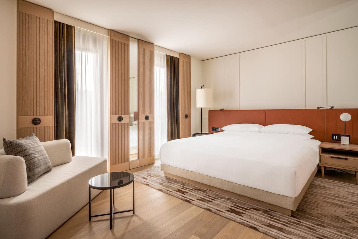 Una habitación de hotel con una cama grande, un sofá, una mesita pequeña y una mesita de noche con lámpara.