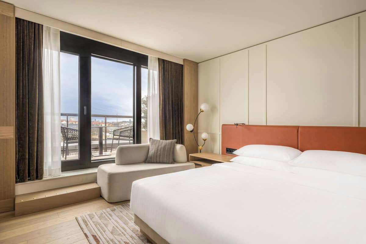 Una habitación de hotel con una cama grande, un sofá, una mesita de noche con lámpara y vistas a la ciudad a través de amplios ventanales.