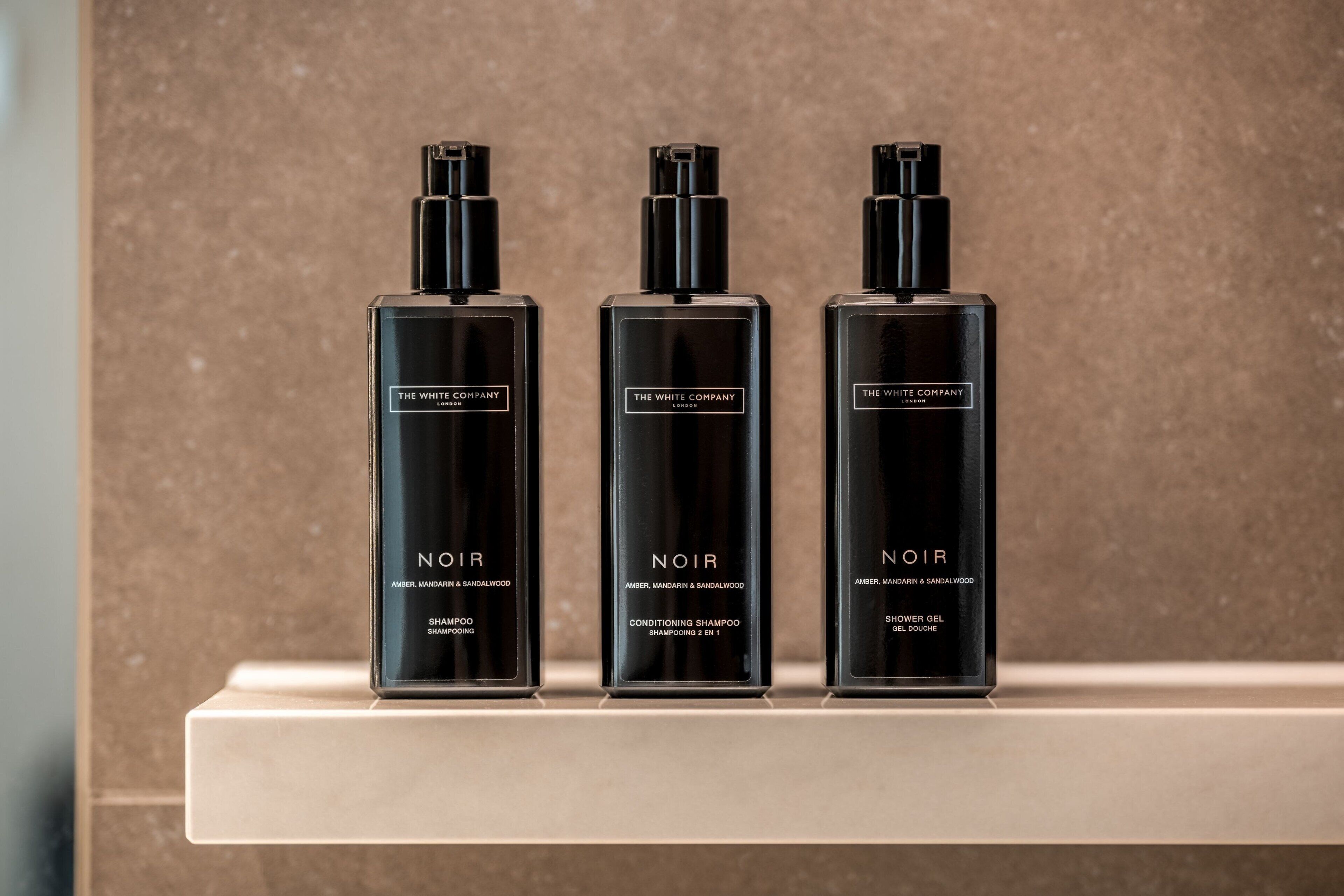 Tres botellas negras de champú, acondicionador y gel de ducha Noir de The White Company sobre un estante blanco.