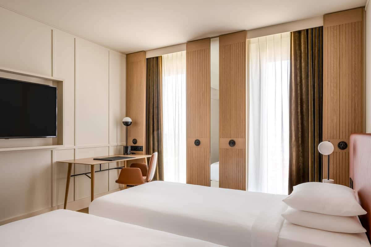 Una habitación de hotel con dos camas, un escritorio, una silla, un televisor y un ventanal grande con cortinas.