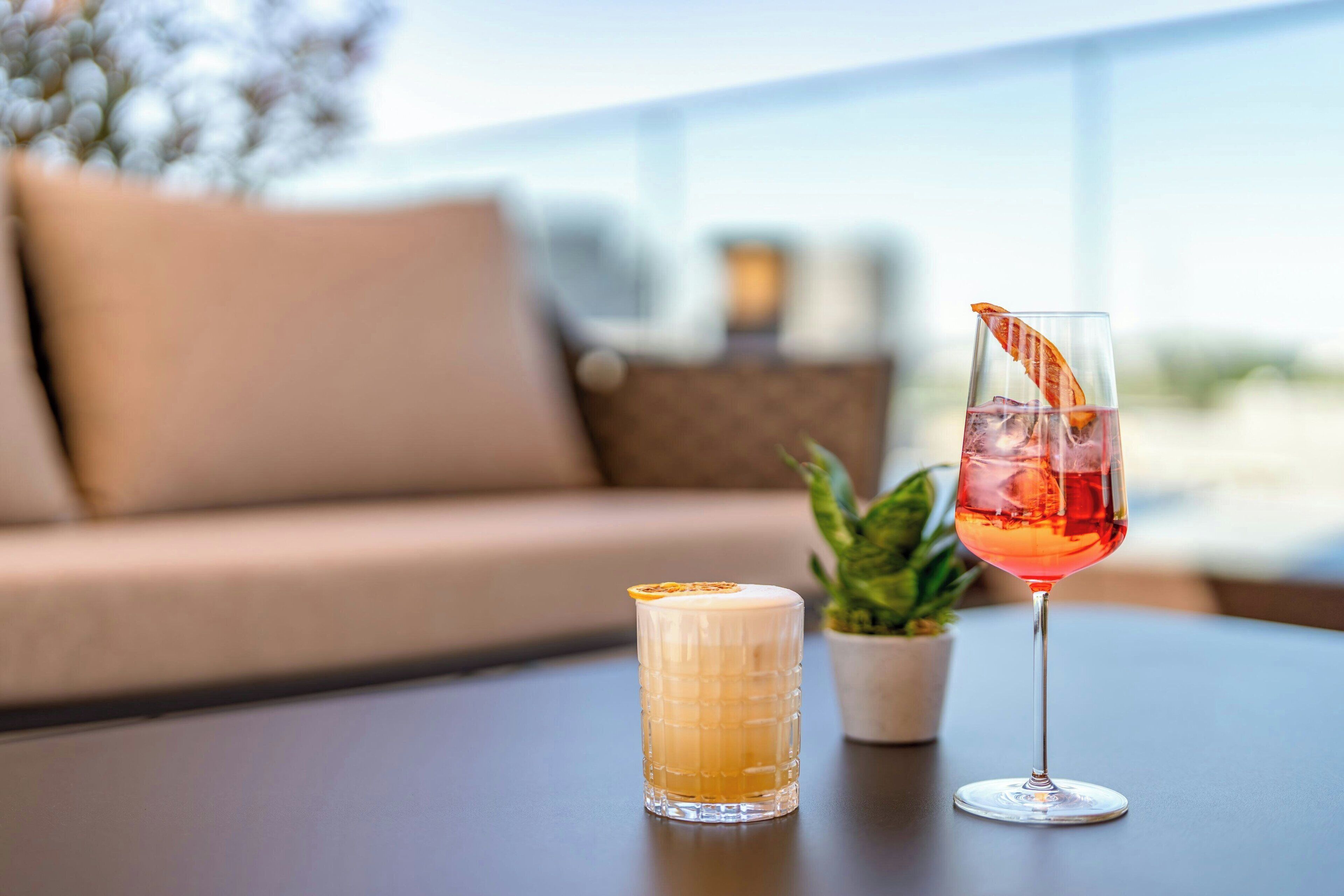 Dos vasos de bebidas sobre una mesa, con un sofá y una planta al fondo.