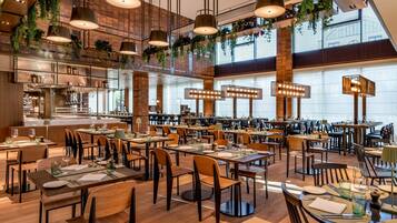 Un restaurante espacioso con varias áreas de comedor, plantas colgantes y grandes ventanales.