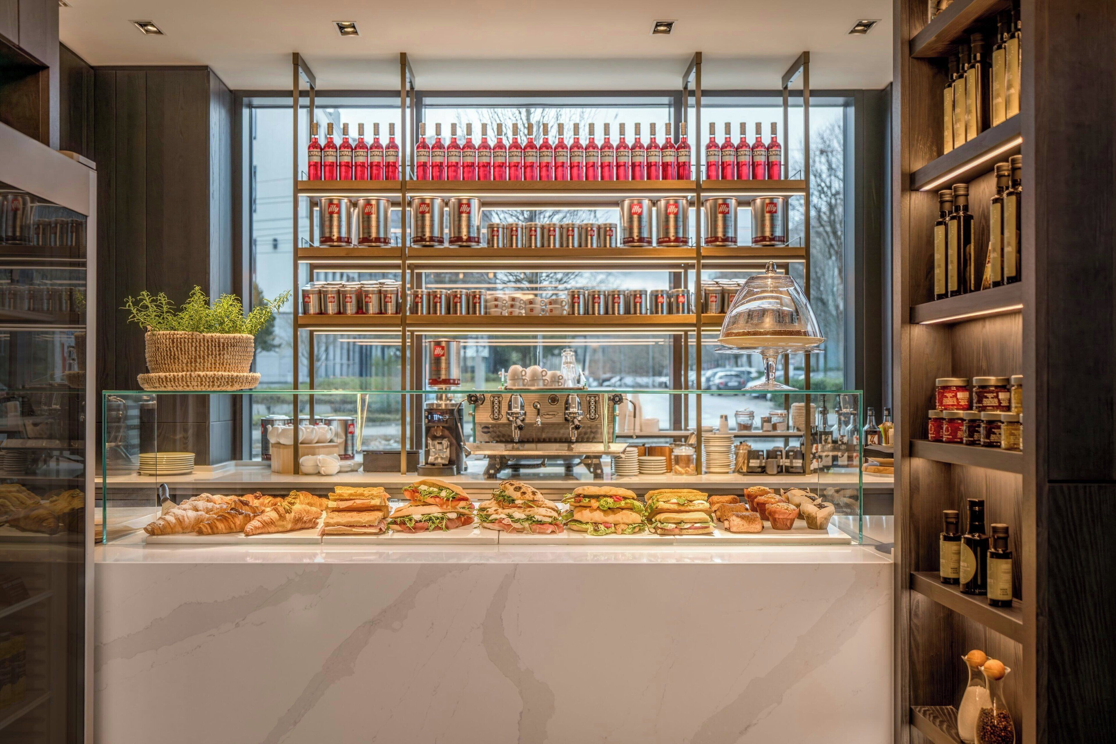 A bakery display with pastries and sandwiches, a coffee machine, and shelves with bottles and jars.