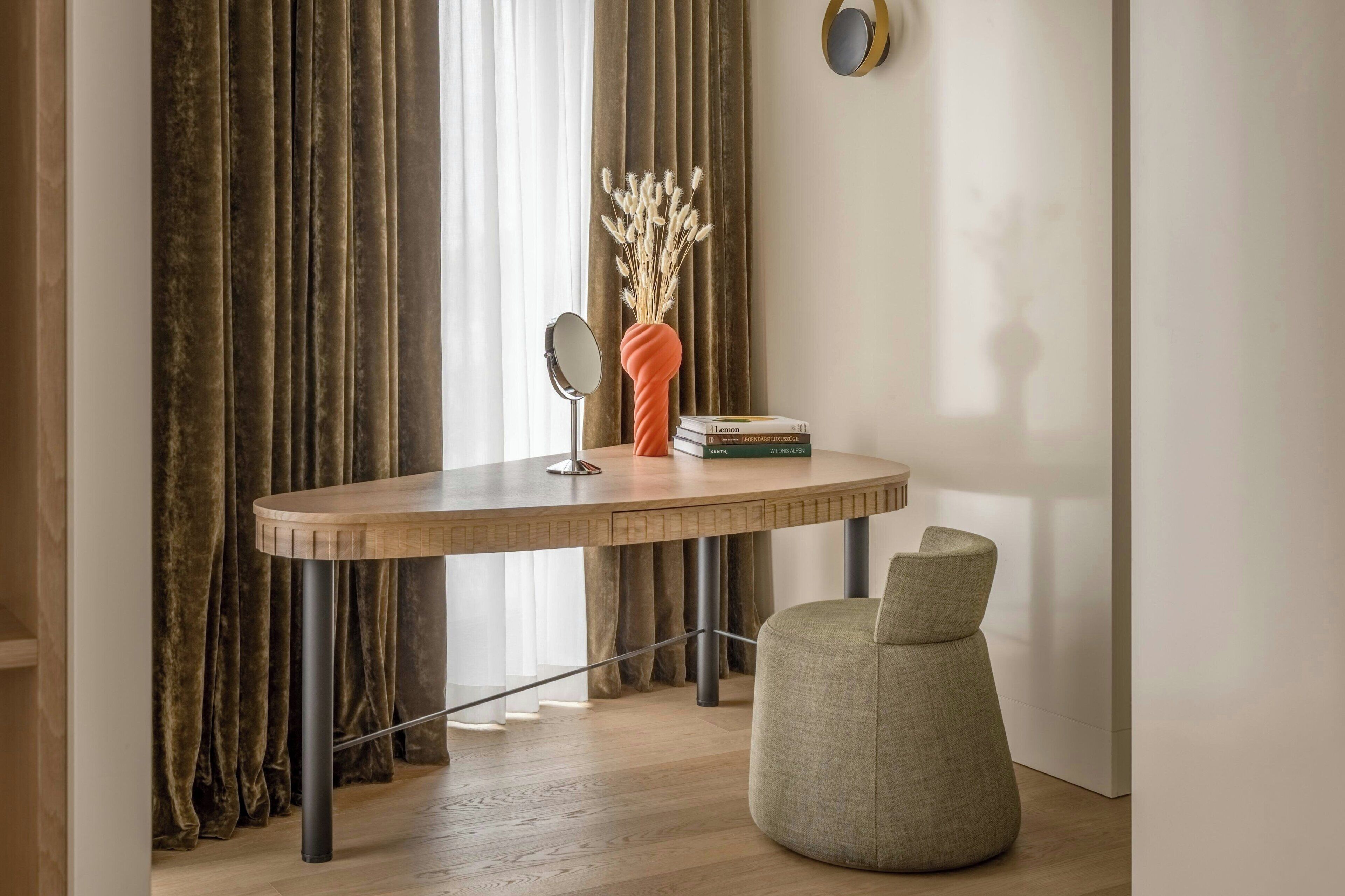 A modern room with a wooden desk, a round mirror, a vase with dried flowers, and a green chair.