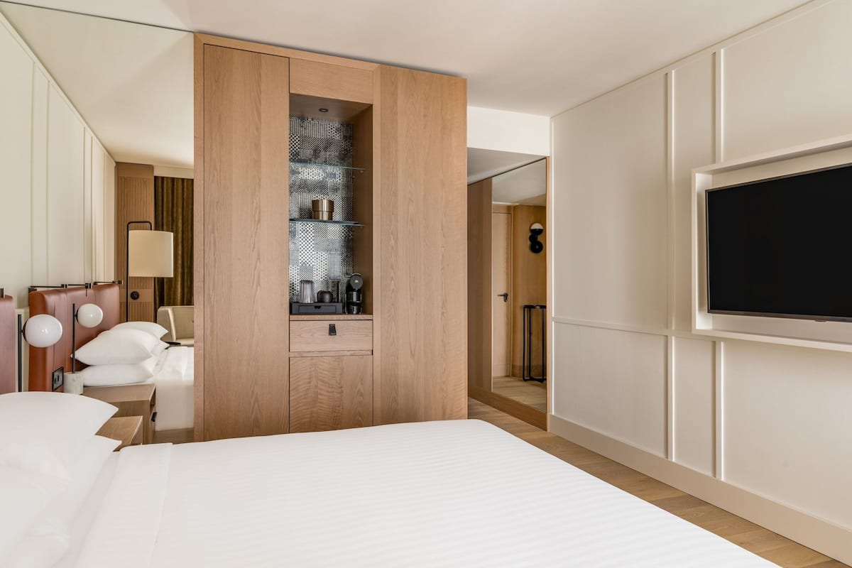 Una habitación de hotel moderna con una cama grande, un armario empotrado, un televisor y un pequeño rincón de descanso con una lámpara.