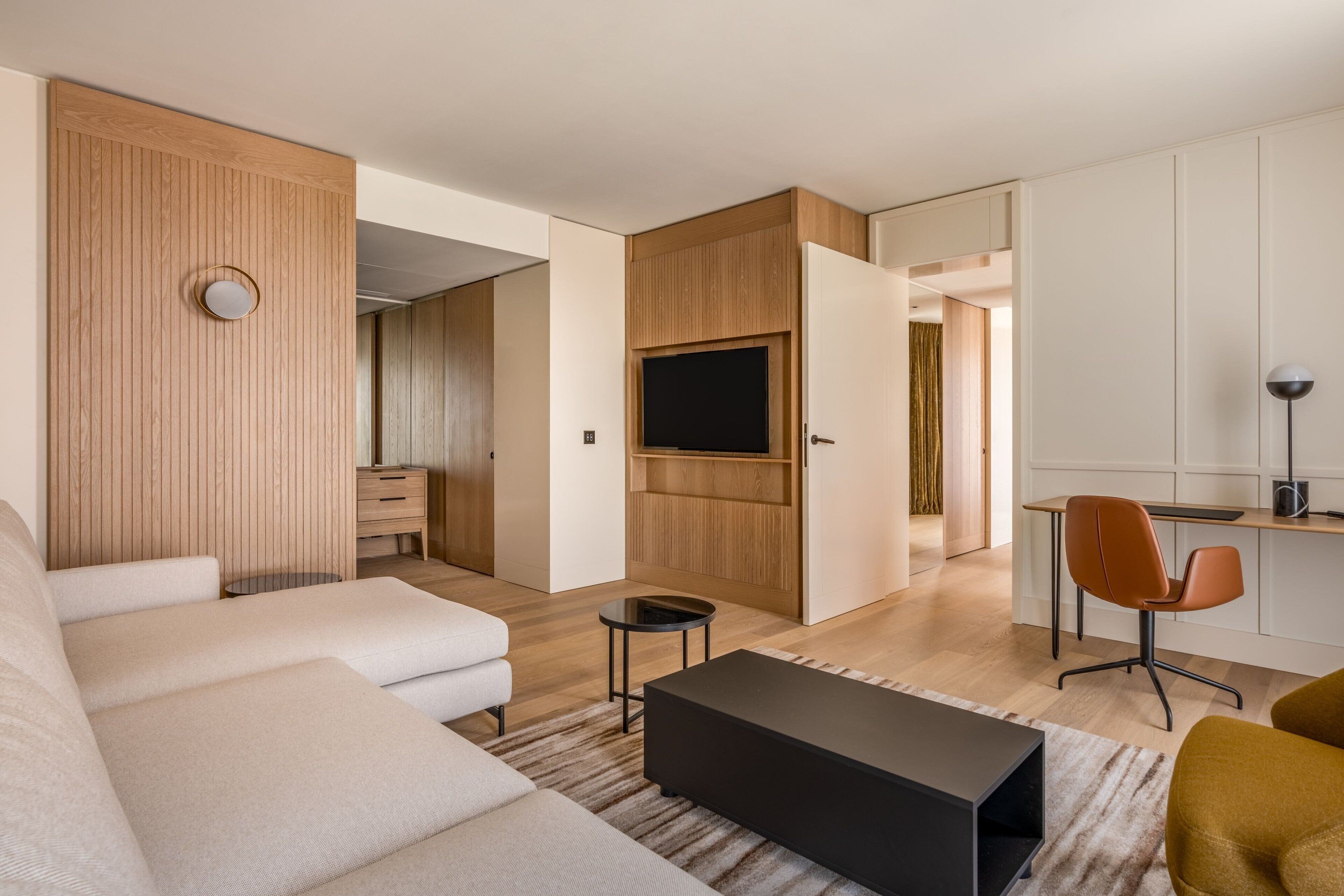 A modern living room with a sofa, a coffee table, and a TV. There is a wooden panel wall and a door leading to another room.