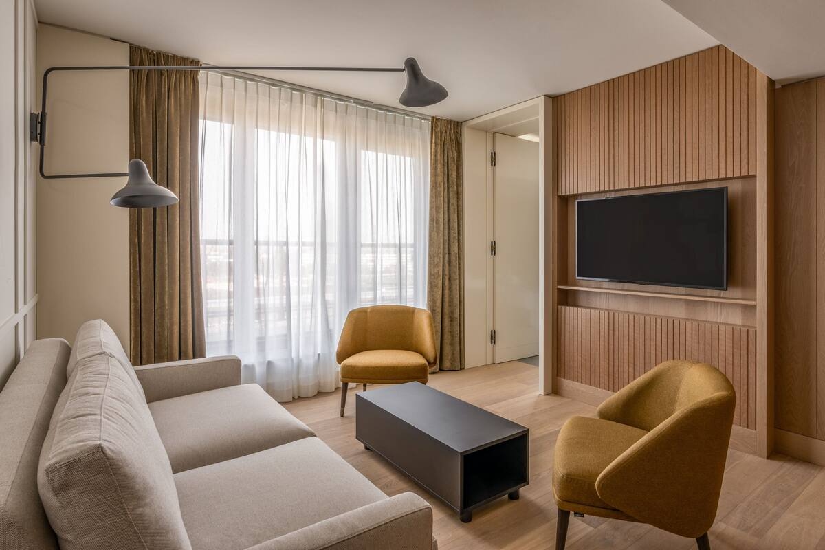 Una sala moderna con un sofá, dos sillones, una mesa de centro, un televisor montado en la pared y un ventanal con cortinas.