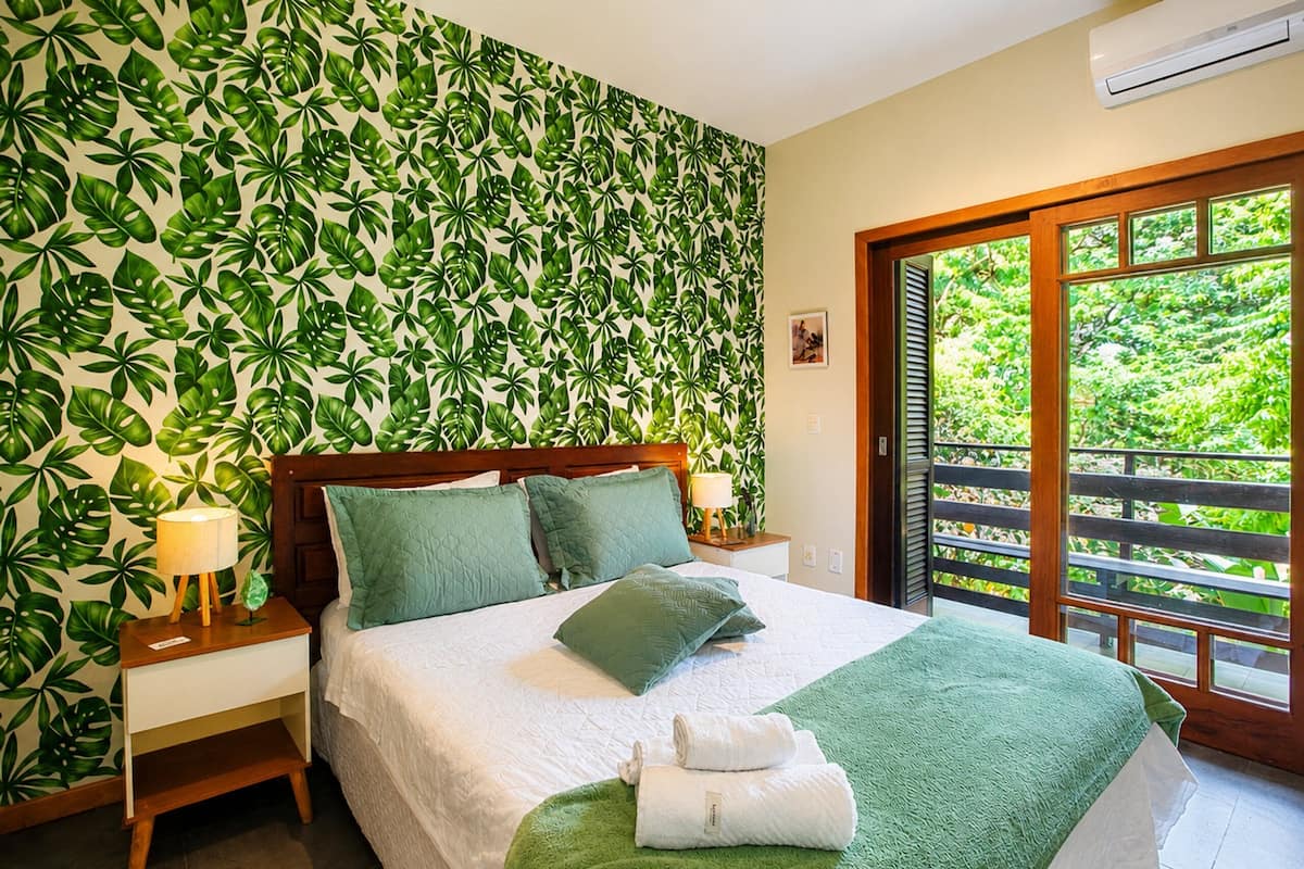 A bedroom with a green leaf-patterned wall, a bed with green bedding, a nightstand with a lamp, and a view of greenery through a sliding door.